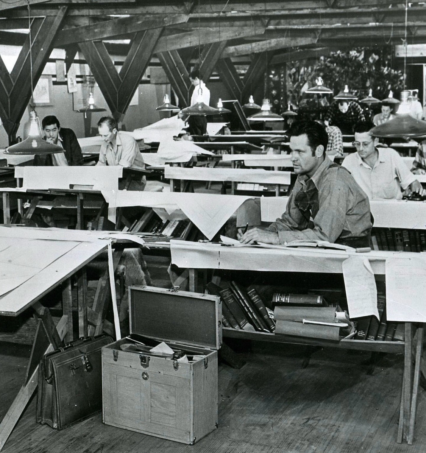 Black and white photograph of a mid-20th-century architectural drafting studio. Several men are seated at large drafting tables with technical drawings, surrounded by wooden beams and overhead lamps. Books and reference materials are stored under the desks. An open toolbox and a leather briefcase sit on the floor in the foreground. The atmosphere is focused and industrious.